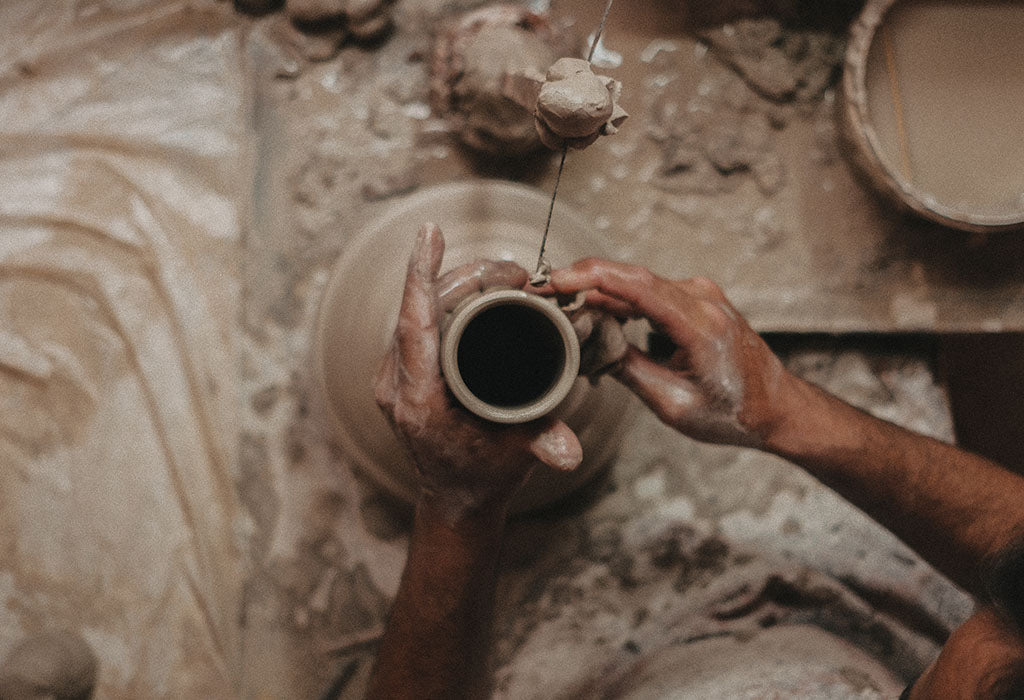 Blick von oben auf zwei Hände, die einen Becher auf einer Töpferscheibe formen. Die Umgebung ist mit Ton bedeckt, ein Drahtwerkzeug trennt den Becher vom Drehteller – Moment der Handarbeit in der Keramikherstellung.