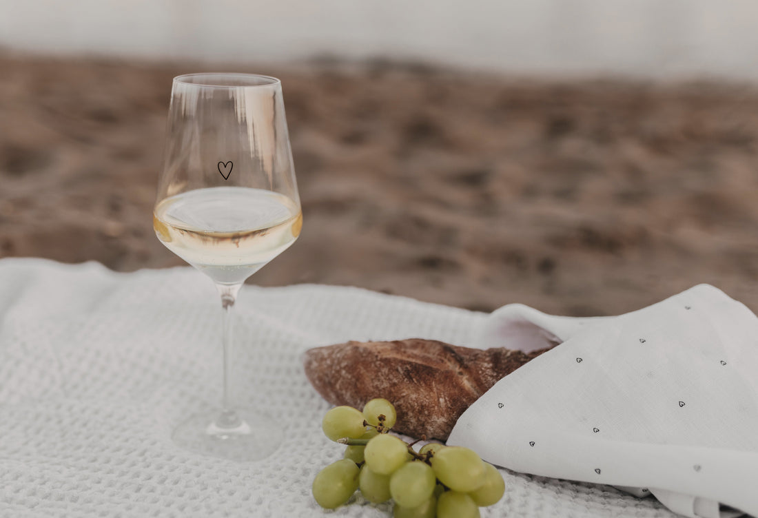 Weinglas mit Herz-Design, dazu Baguette und Trauben auf Picknickdecke am Strand.