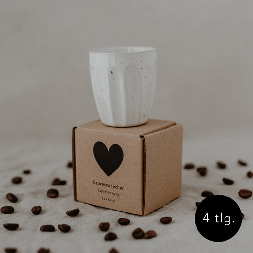 Espressobecher komplett lasiert mit braunen Sprenklern steht auf einem Umkarton mit einem schwarzen Herzen. Im Hintergrund liegen viele Kaffeebohnen.