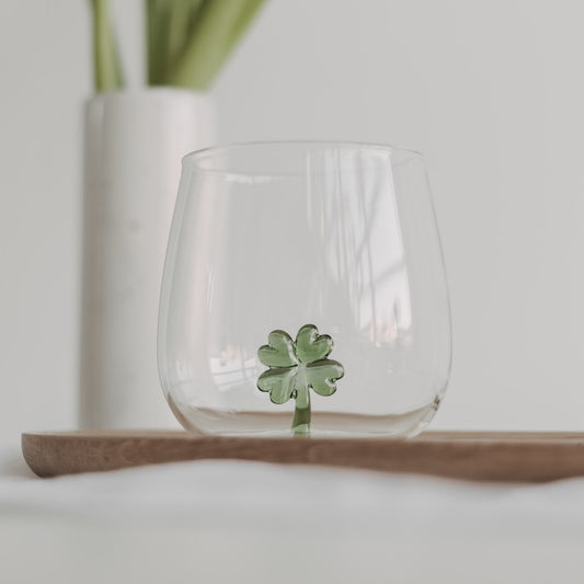Trinkglas mit einer grünen Kleeblattfigur im Glasboden, auf einem Holztablett platziert, heller Hintergrund.