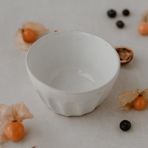 Eine Schüssel mit weißer Lasur und Sprenkeln steht auf einem beigen Hintergrund. Blaubeeren, Wallnüsse und Physalis liegen dahinter.