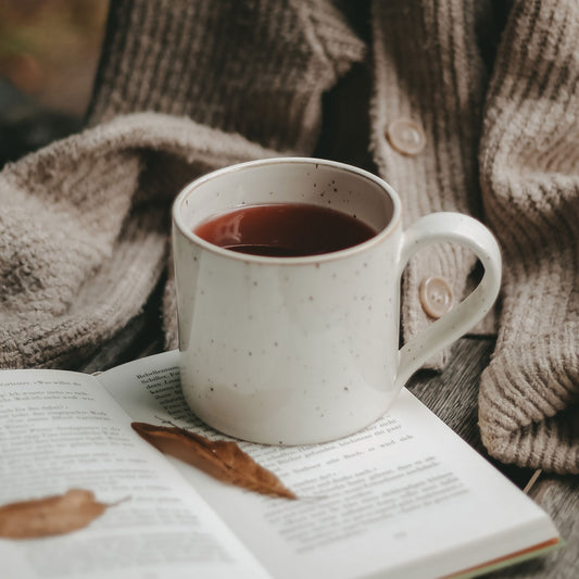 Weiße, gesprenkelte Keramiktasse mit Henkel, gefüllt mit dunklem Tee, steht auf einem offenen Buch. Daneben liegt ein herbstliches Blatt, im Hintergrund ist eine grob gestrickte, beigefarbene Strickjacke zu sehen. Das Bild vermittelt eine warme, ruhige Herbststimmung – perfekt für entspannte Teemomente.
