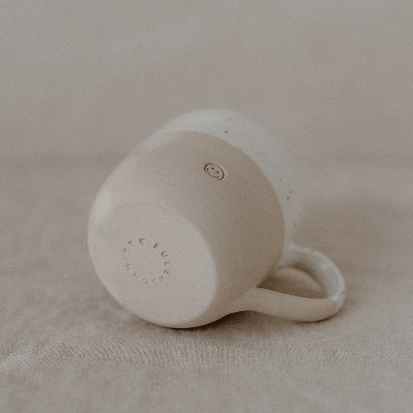 Keramiktasse in Beige und Weiß liegt auf der Seite, Henkel nach rechts gerichtet. Sichtbar ist die Unterseite mit eingeprägtem Eulenschnitt-Schriftzug sowie das kleine Smiley-Symbol auf der Vorderseite. Die Tasse liegt auf hellem Leinenstoff – detailreiche Aufnahme zur Präsentation von Markenlogo und Handwerksstruktur.