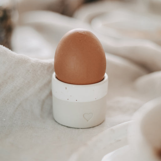 Braunes Ei in einem naturfarbenen Keramik-Eierbecher mit glasiertem weißen Rand und Herzprägung, arrangiert auf weichem Leinentuch.