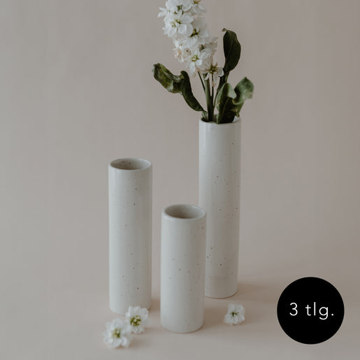 Drei zylindrische, schlanke Keramikvasen in verschiedenen Höhen, in natürlichem Cremeweiß mit feinen Sprenkeln. Eine Vase ist mit weißen Blumen bestückt, die anderen zwei sind leer. Daneben liegen locker weiße Blüten, rechts unten ein schwarzer Kreis mit der Aufschrift „3 tlg.“.