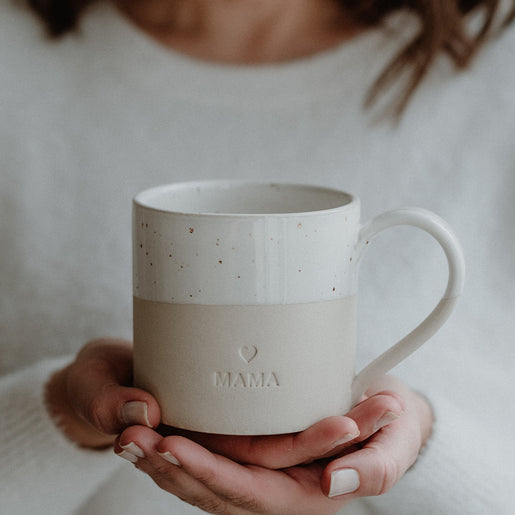 Zwei Hände halten eine beige-weiße Keramiktasse mit gesprenkelter Glasur und Henkel. Auf der Vorderseite ist ein kleines Herz sowie das Wort ‚MAMA‘ eingeprägt. Die Person trägt einen weichen, weißen Pullover – die Szene strahlt Geborgenheit und Dankbarkeit aus, ideal als Geschenk für Mütter.