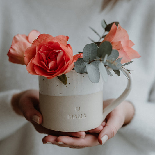 Zwei Hände halten eine beige-weiße Keramiktasse mit gesprenkelter Glasur und der Gravur ‚MAMA‘ unter einem kleinen Herzsymbol. Die Tasse ist mit rosafarbenen Rosen und Eukalyptuszweigen gefüllt. Der weiche Hintergrund und die sanfte Farbgebung betonen Zärtlichkeit und Wertschätzung – ideal als Geschenk für Mütter.