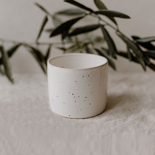 Schlichter, zylindrischer Keramikbecher mit feiner brauner Sprenkelung auf hellem Grund. Im Hintergrund sind Olivenzweige stilvoll arrangiert.
