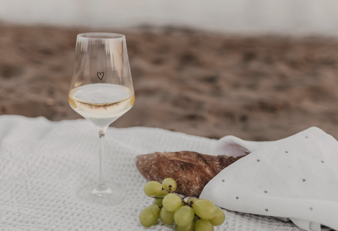 Weinglas mit Herz-Design, dazu Baguette und Trauben auf Picknickdecke am Strand.