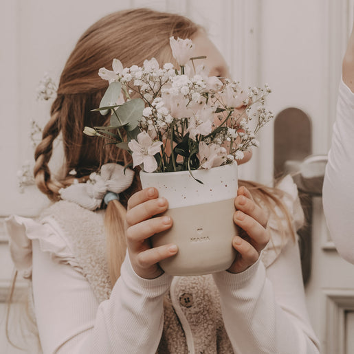 Ein Becher mit der Prägung "Mama" wird von einem süßen Mädchen mit einem geflochtenem Zopf gehalten.