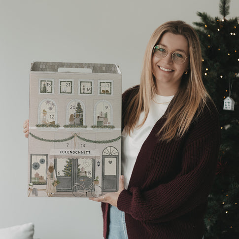 Frau präsentiert den Eulenschnitt Signature-Adventskalender neben einem beleuchteten Weihnachtsbaum.