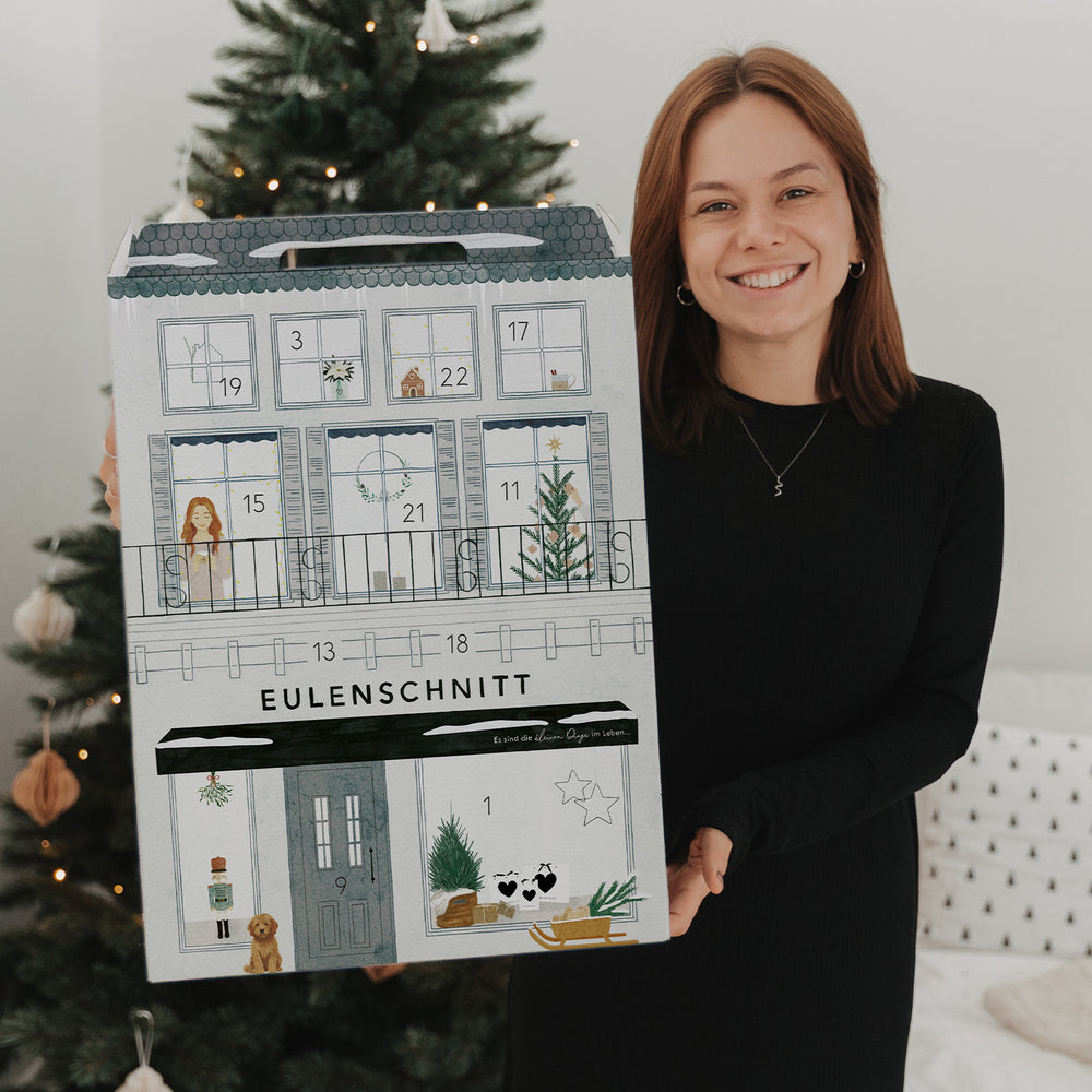 Frau mit Eulenschnitt Adventskalender vor festlich geschmücktem Weihnachtsbaum in gemütlicher Atmosphäre.