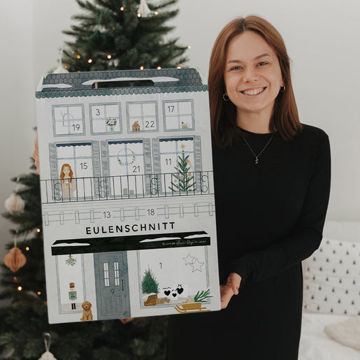 Frau mit Eulenschnitt Adventskalender vor festlich geschmücktem Weihnachtsbaum in gemütlicher Atmosphäre.
