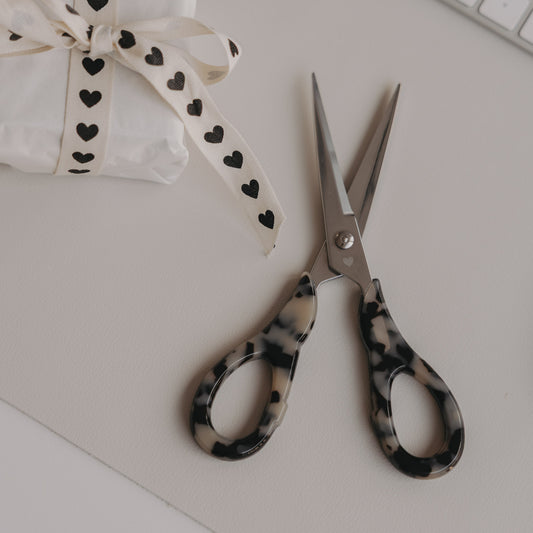 Silberne Schere mit Leo-Griff neben Geschenk mit Herzband auf hellem Hintergrund