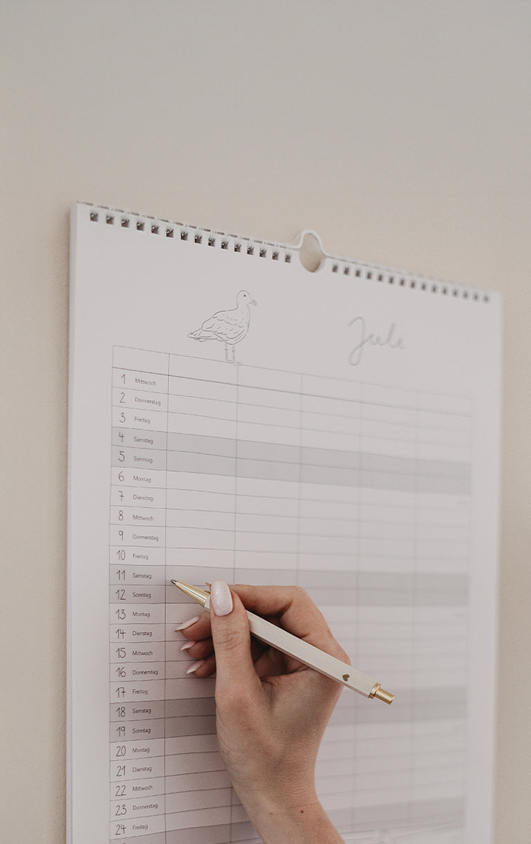 Familienkalender 2026 mit liebevoll gestalteten Illustrationen hängt an der Wand, jemand trägt Termine mit unserem cremefarbenen Kugelschreiber ein. Oben im Bild steht: „Kalender – für all die schönen Momente“ und unten als Schaltfläche „Zu den Kalendern“