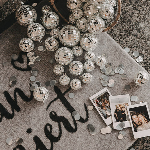 Graue Fußmatte mit schwarzem Schriftzug „Hereinspaziert“, dekoriert mit silbernen Discokugeln, Konfetti und Polaroid-Fotos.