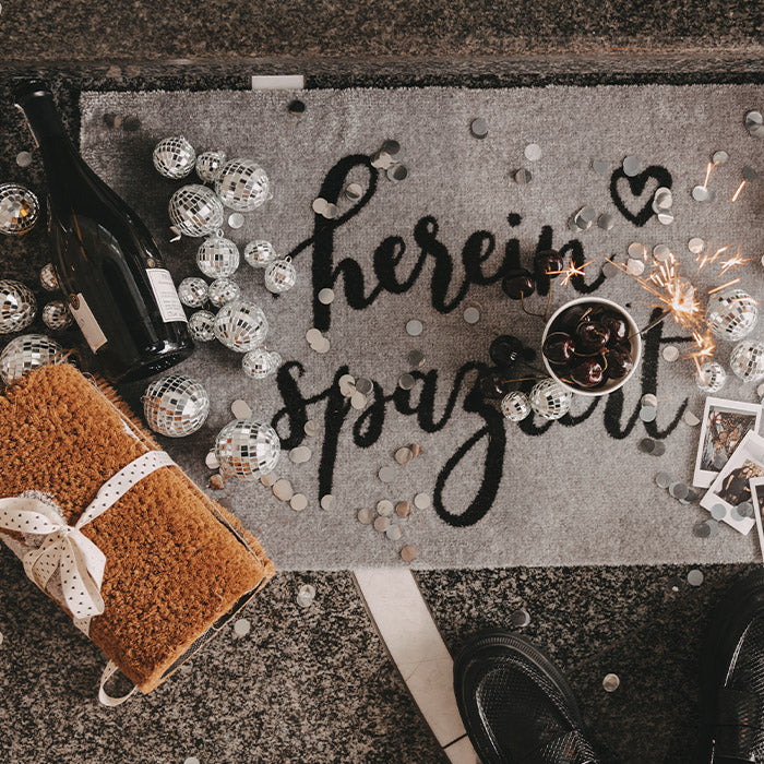 Graue Fußmatte mit schwarzem Schriftzug „Hereinspaziert“ in einer festlichen Geburtstagsszene mit Discokugeln, Konfetti, Wunderkerzen, Getränken und einem verpackten Geschenk.
