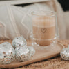 Doppelwandiges Cappuccino-Glas mit Herzmotiv auf einem Holzbrett neben kleinen verspiegelten Dekokugeln