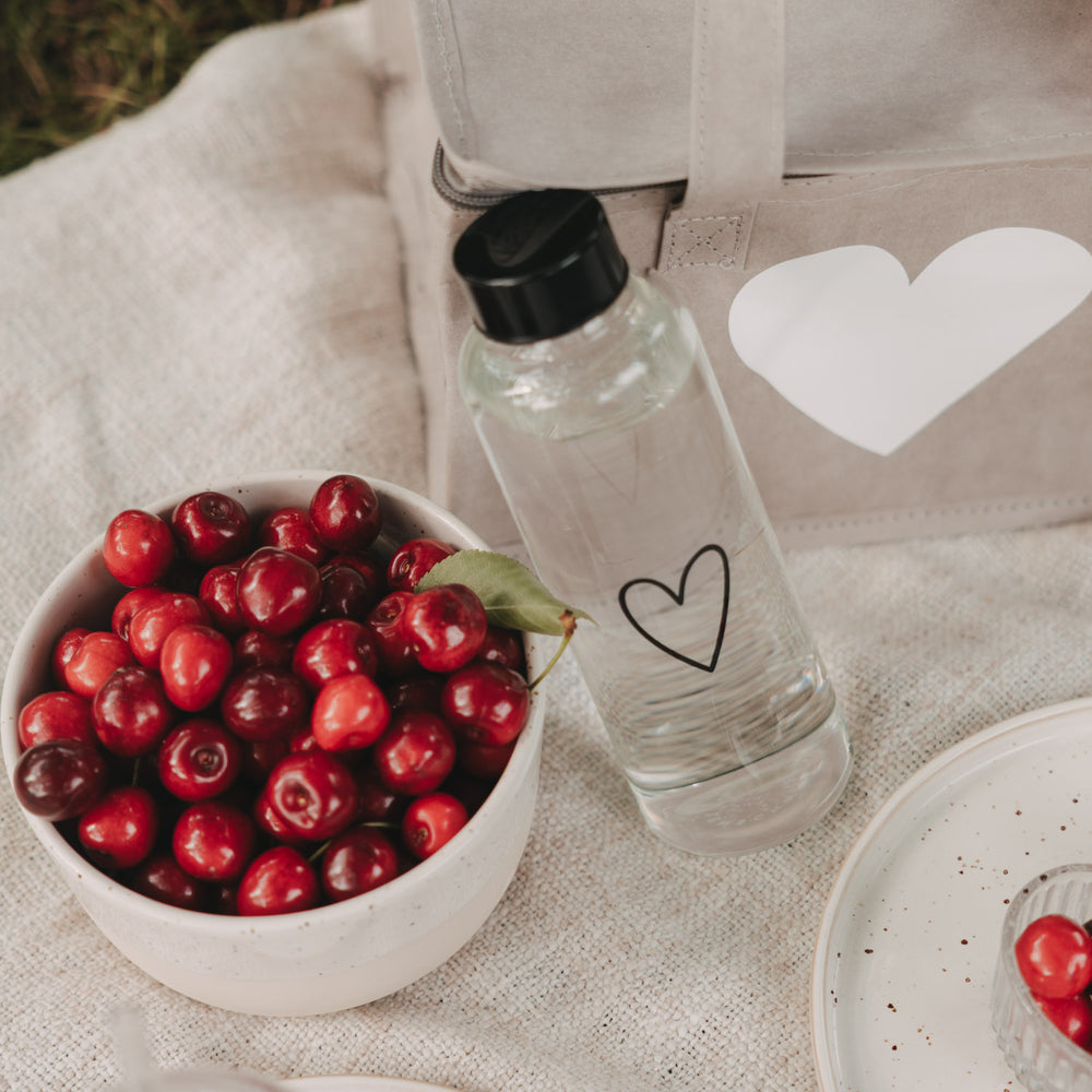 Glas-Trinkflasche mit schwarzem Herz neben Schale mit Kirschen und Kühltasche beim Picknick