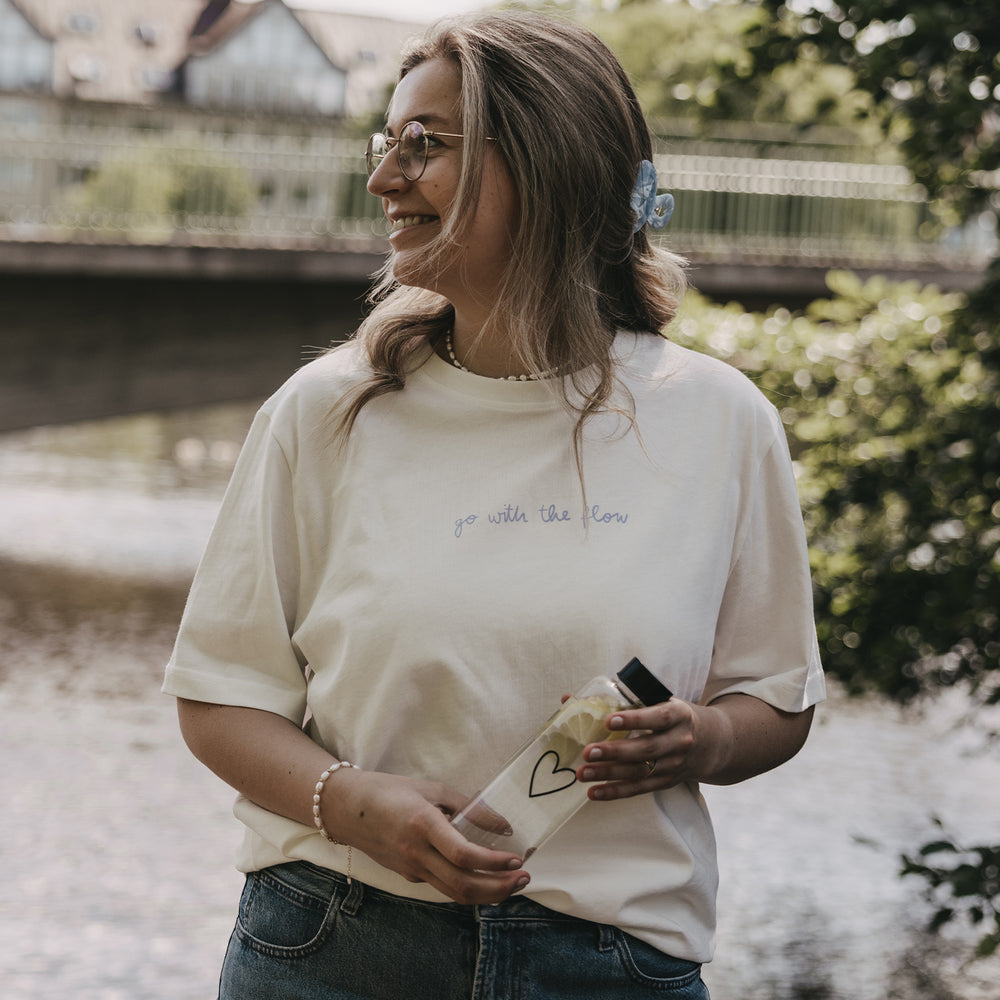 Lächelnde Frau mit cremefarbenem T-Shirt und transparenter Trinkflasche mit Herzmotiv am Flussufer.