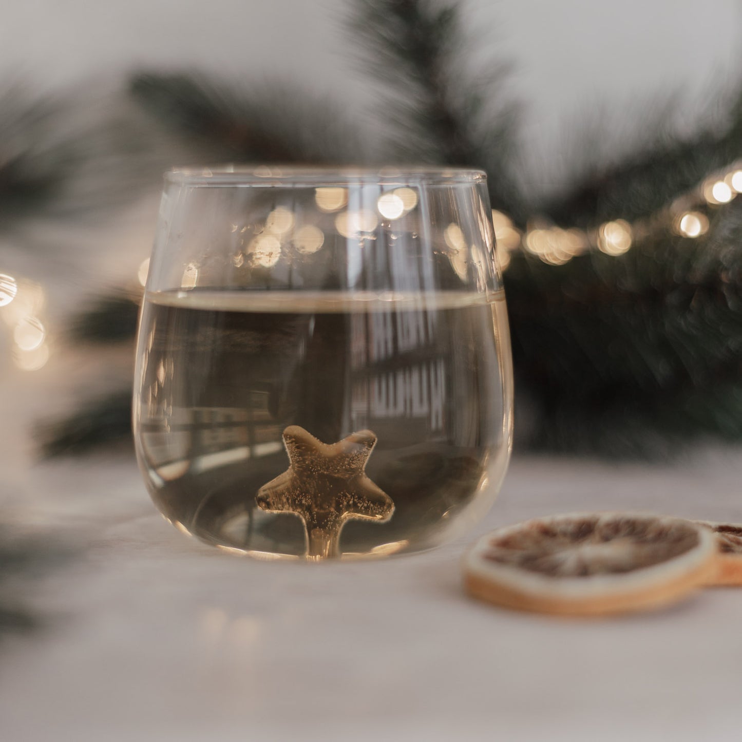 Nahaufnahme Glas mit goldfarbenem Stern im Boden, weihnachtliche Atmosphäre mit Lichterkette und Orangenscheiben