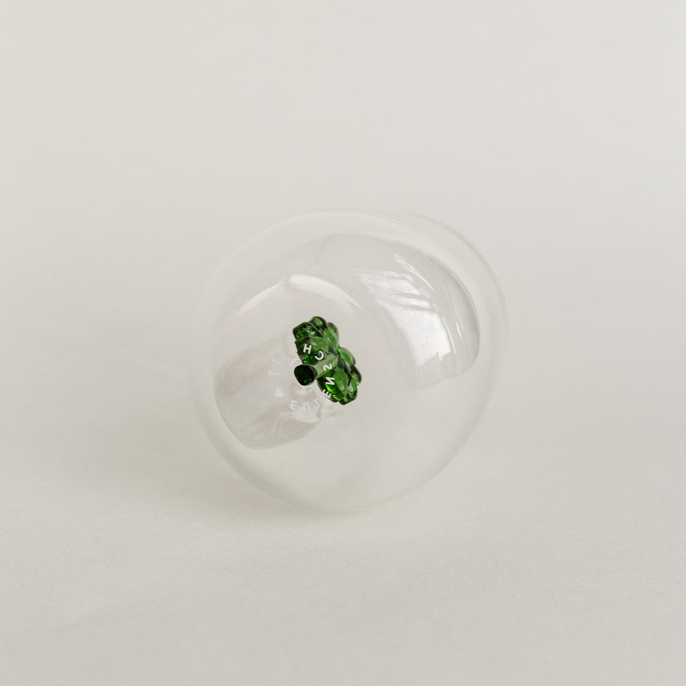 Figurglas mit grünem Kleeblatt im Inneren, transparentes Glas mit Eulenschnitt Logo auf hellem Hintergrund von unten fotografiert.