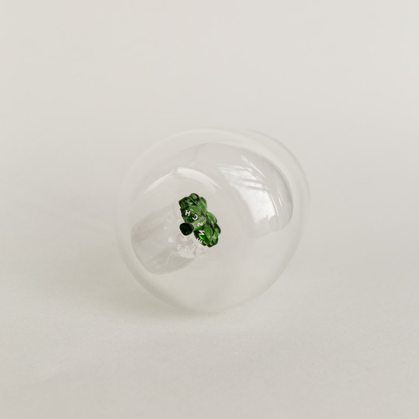 Figurglas mit grünem Kleeblatt im Inneren, transparentes Glas mit Eulenschnitt Logo auf hellem Hintergrund von unten fotografiert.