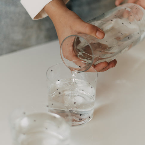 Wasser wird aus einer Glaskaraffe mit kleinen schwarzen Herzen in ein passendes Trinkglas gegossen