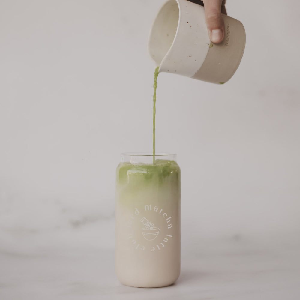 Matcha wird aus einem kleinen Krug in ein Glas mit eisgekühltem Latte gegossen. Das minimalistische Glas steht vor einem hellen, ruhigen Hintergrund.