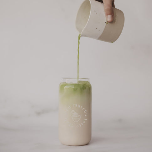 Matcha wird aus einem kleinen Krug in ein Glas mit eisgekühltem Latte gegossen. Das minimalistische Glas steht vor einem hellen, ruhigen Hintergrund.