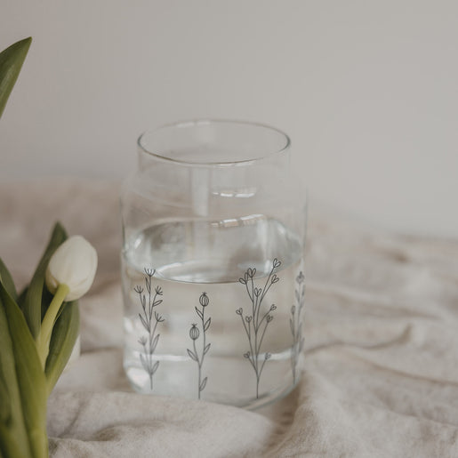 Glasvase mit Blumenwiesen-Motiv gefüllt mit Wasser auf hellem Stoff mit Tulpe daneben