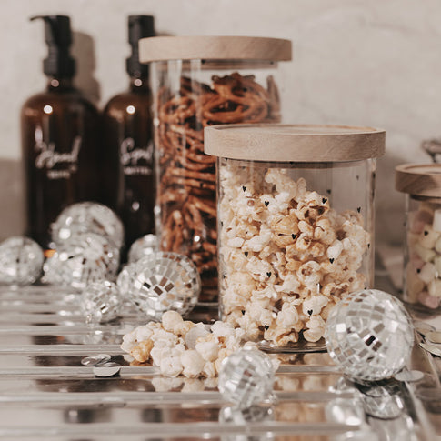 Glasbehälter mit kleinen schwarzen Herzen, gefüllt mit Popcorn und mit Holzdeckel, dekoriert mit silbernen Discokugeln.
