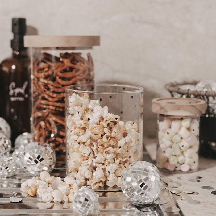 Herzchen-Glas gefüllt mit Popcorn neben Vorratsgläsern mit Brezeln und Marshmallows, dekoriert mit silbernen Discokugeln und Konfetti.