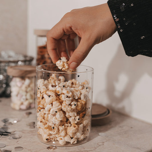 Hand greift nach Popcorn aus einem Glasbehälter mit kleinen schwarzen Herzen.