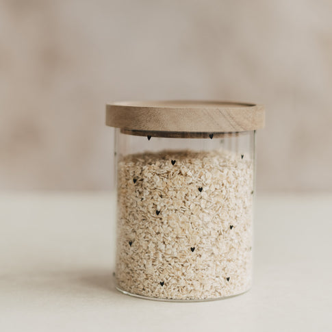 Vorratsglas mit schwarzen Herzen und Holzdeckel, gefüllt mit Haferflocken, vor hellem Hintergrund.