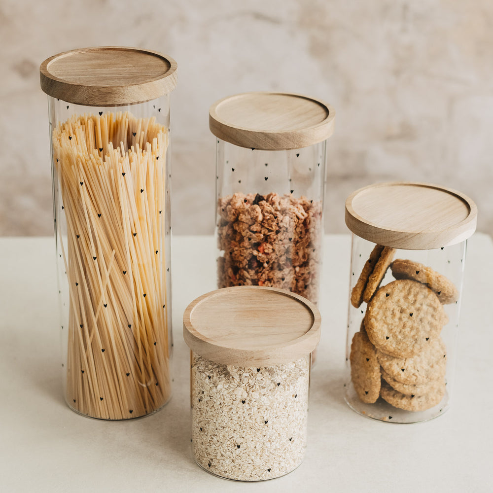 Vier unterschiedlich große Vorratsgläser mit schwarzen Herzchen und Holzdeckeln, gefüllt mit verschiedenen Lebensmitteln.