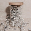 Hoher Glasbehälter mit kleinen schwarzen Herzen und Holzdeckel, gefüllt mit silbernen Discokugeln und umgeben von weiteren Discokugeln und Konfetti.