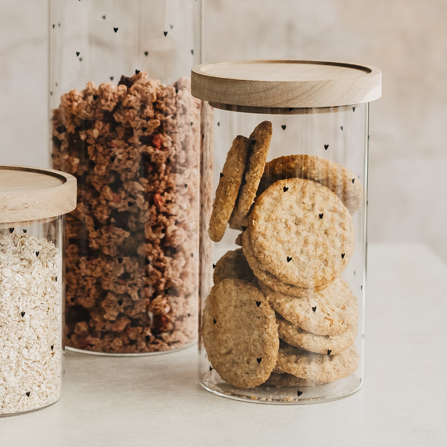 Vorratsglas mit schwarzen Herzchen und Holzdeckel, gefüllt mit Keksen, neben Gläsern mit Müsli und Haferflocken.