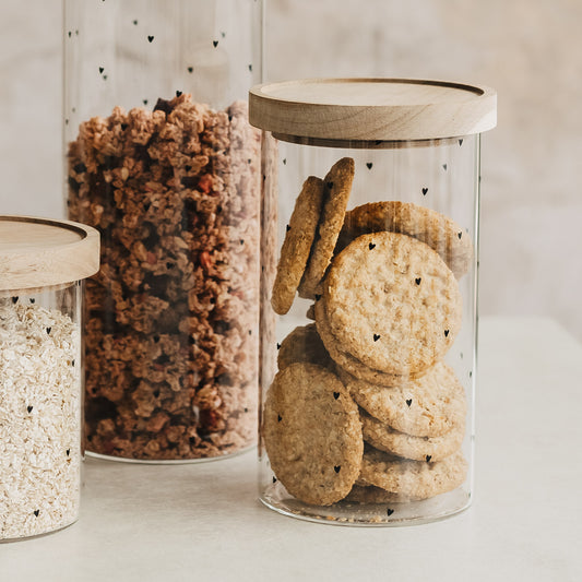 Vorratsglas mit schwarzen Herzchen und Holzdeckel, gefüllt mit Keksen, neben Gläsern mit Müsli und Haferflocken.