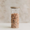 Hohes Vorratsglas mit kleinen schwarzen Herzen, Holzdeckel und Müsli auf hellem Hintergrund.