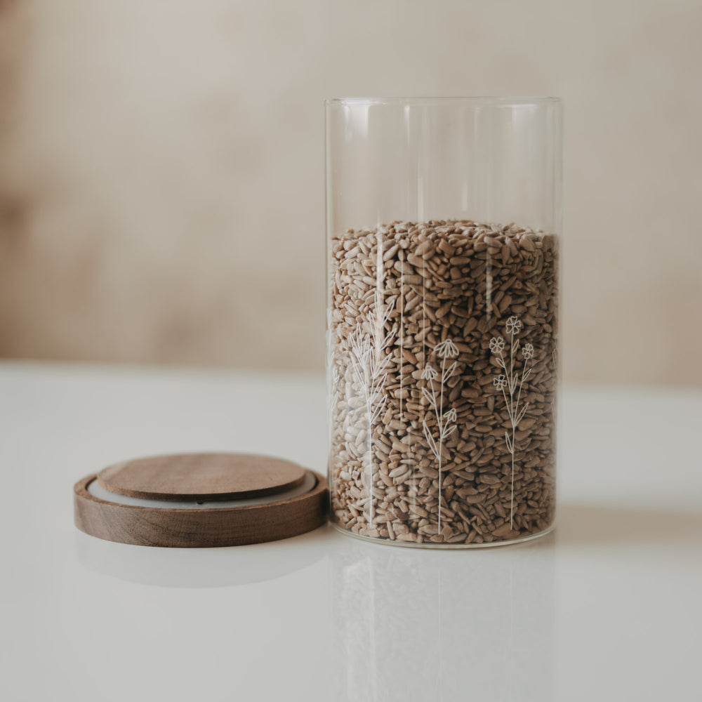 Geöffneter Glas-Vorratsbehälter mit weißem Wildblumen-Motiv, gefüllt mit Sonnenblumenkernen, daneben der Holzdeckel