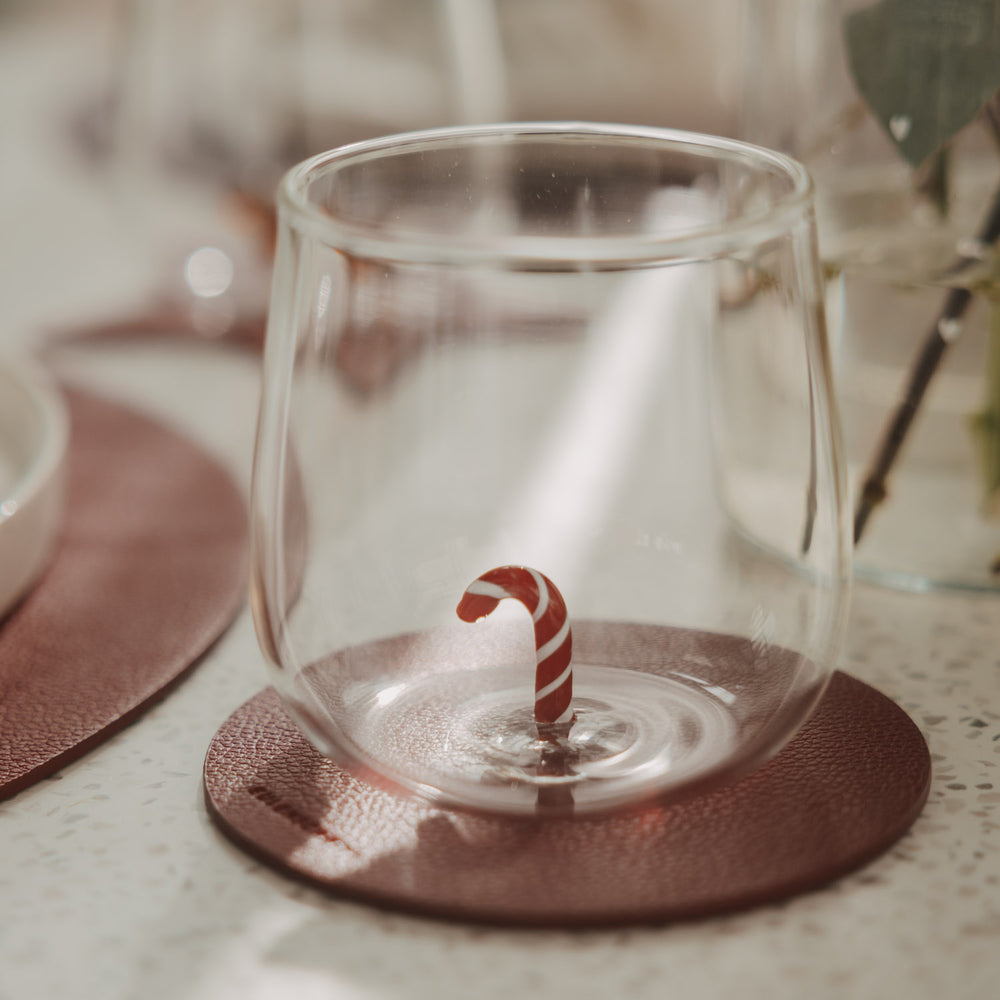Transparentes Figurenglas mit rot-weißer Zuckerstange im Glasboden auf einem braunen Untersetzer.