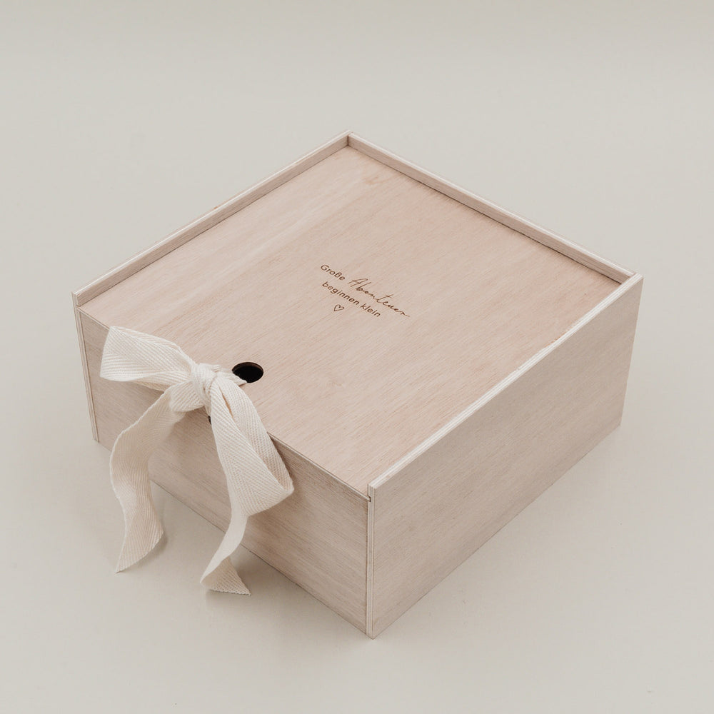 Geschlossene Holzbox mit Gravur „Große Abenteuer beginnen klein“ und cremefarbener Schleife.