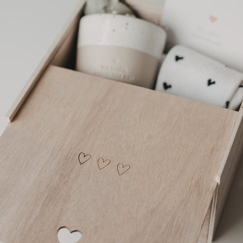 Detailaufnahme der halb geöffneten Erinnerungsbox mit drei eingravierten Herzen auf dem Holzdeckel. Im Inneren sind ein Keramikbecher, eine Karte und gemusterte Socken zu erkennen.