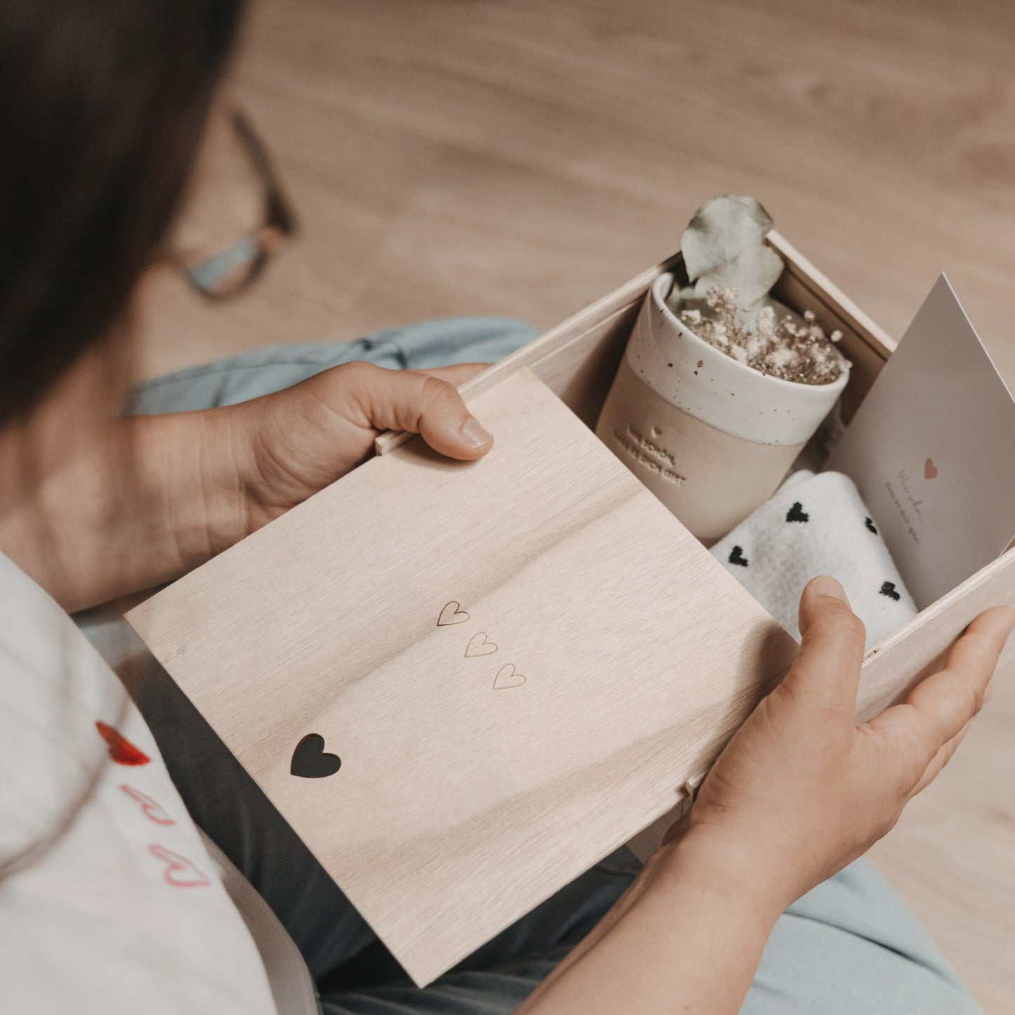Person hält eine geöffnete Erinnerungsbox aus Holz in den Händen. Im Inneren befinden sich ein Keramikbecher, Trockenblumen, eine Karte und weiche Socken mit Herzmuster – liebevoll arrangiert als Geschenkset.
