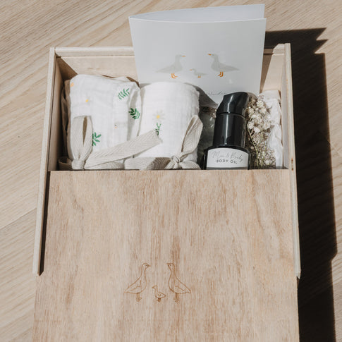 Geöffnete Erinnerungsbox aus Holz mit gravierter Gänsefamilie auf dem Schiebedeckel. Die Box ist mit Musselintüchern, einer Grußkarte, Trockenblumen und einem Pflegeöl gefüllt und eignet sich ideal als Geschenk zur Geburt oder Babyparty.