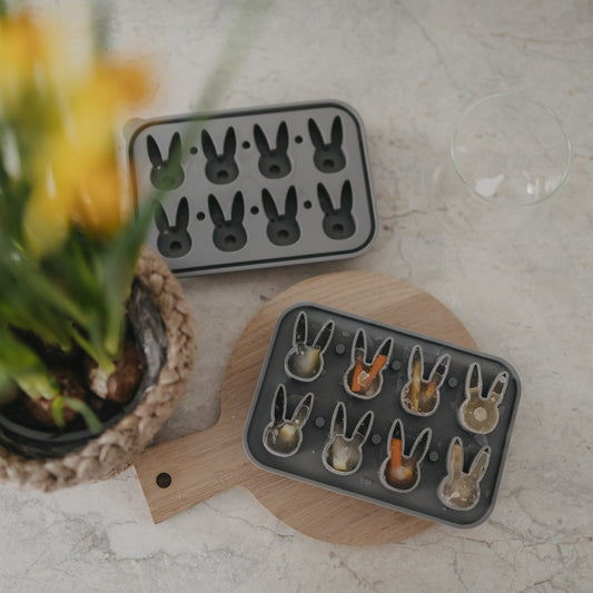 Ansicht von oben der grauen Eiswürfelform aus Silikon mit erhöhten Hasenformen gefüllt mit Eis und Orangen- und Zitronenscheiben, dekoriert auf einem runden Holzbrett. Daneben steht eine Pflanze.
