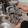Graue Silikon-Eiswürfelform für sechs herzförmige Eiswürfel, gefüllt mit gefrorenen Früchten und Kräutern, wird in den Händen gehalten.