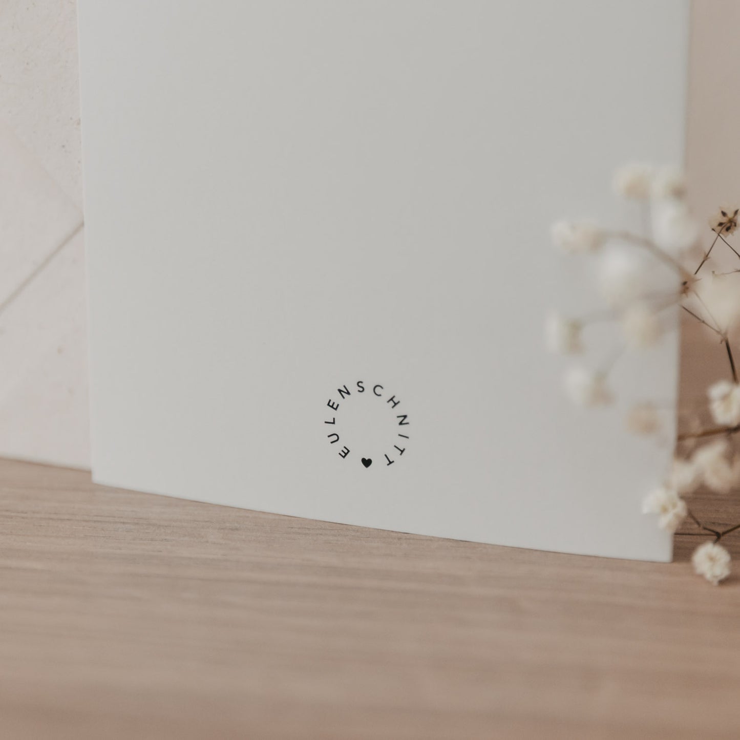 Weiße Klappkarte mit dem Schriftzug 'Eulenschnitt' als Logo, auf Holztisch neben getrockneten Blüten, Hochzeitskarte mit Motiv Hochzeitsauto