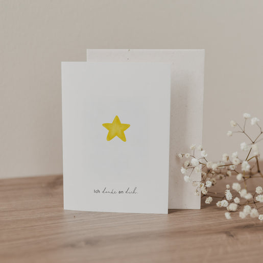 Klappkarte mit gelbem Stern und Schrift „Ich denke an dich“, stehend mit Umschlag auf Holzoberfläche dekoriert mit Trockenblumen.